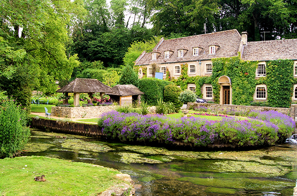 A picturesque stone cottage surrounded by lush greenery and vibrant purple flowers near a tranquil stream. The property features a gazebo and serene garden, creating an idyllic countryside scene.