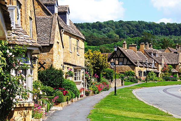 A picturesque village street lined with charming stone cottages, vibrant flowers, and lush greenery against a backdrop of rolling hills.