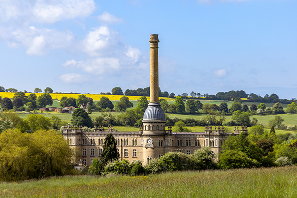 A historic building with a tall chimney, surrounded by lush green fields and trees under a blue sky. The architecture features a large dome and intricate details. Ideal for those interested in heritage sites.