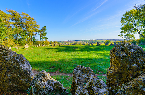 A scenic landscape featuring large stones in the foreground, lush green grass, and a clear blue sky, with distant fields and trees in the background, capturing a peaceful outdoor environment.