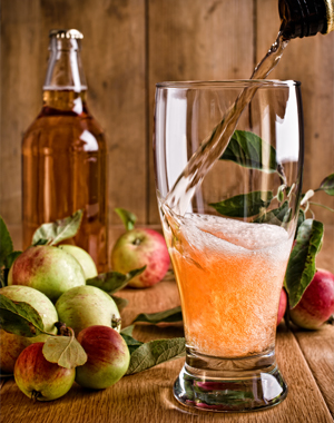 Bottle of cider being poured into glass with background of ripe apples and cider bottles