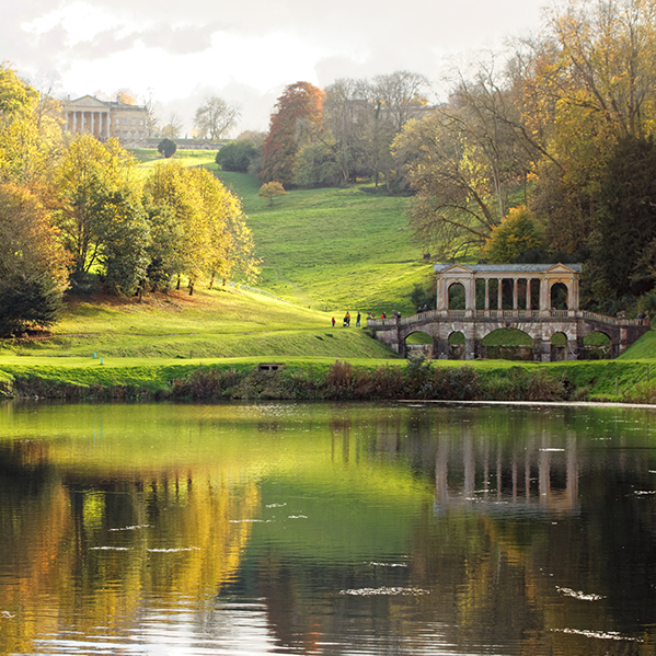 Scenic view of a park with lush green hills, colorful autumn trees, and a reflective lake, featuring a classic stone bridge and a grand historic building in the background.