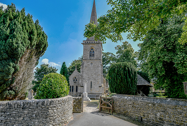 A picturesque church surrounded by lush greenery, featuring a tall spire and stone walls, with pathways leading through the cemetery and trees shading the entrance.