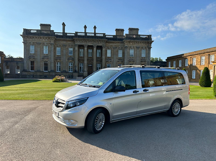 Silver van parked in front of an impressive historic building with manicured lawns and clear blue skies, symbolizing luxury travel.